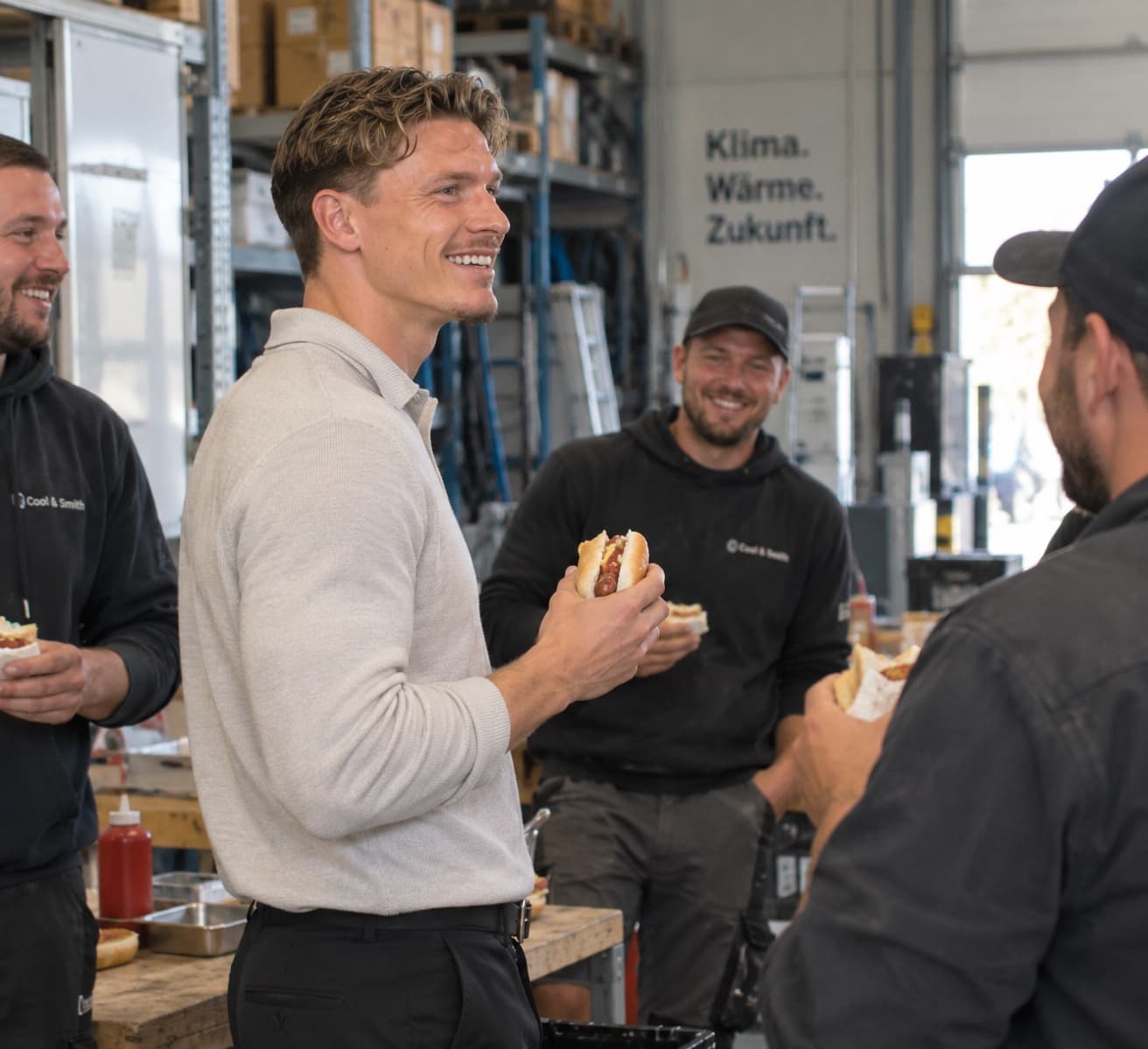 Luca Christ steht morgens mit vier Monteuren im Lager eines Handwerksbetriebs, alle essen eine Wurst im Brötchen